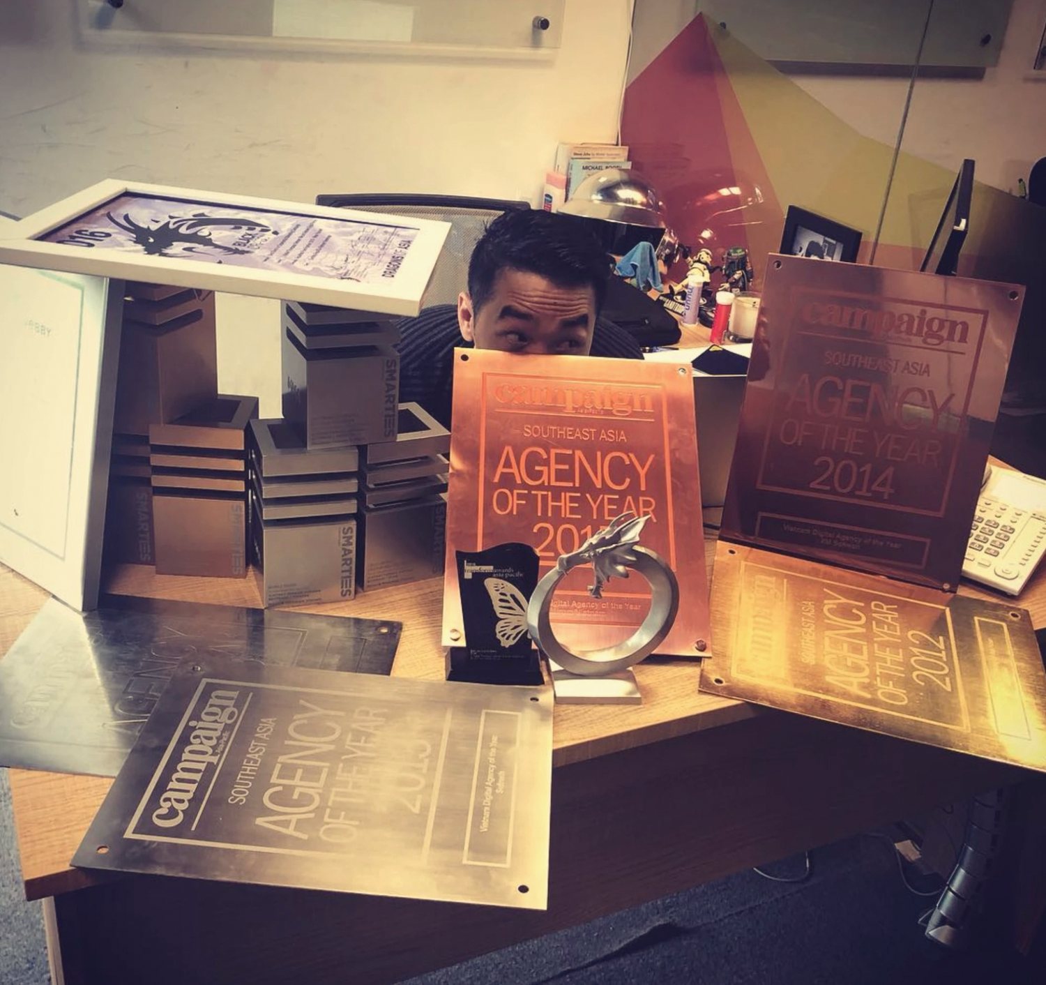 Ly peeking over a desk covered in Agency of the Year plaques and trophies